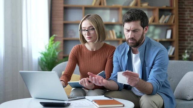 Confused married couple having difficulty doing household finances using application on laptop computer sitting at home office. Puzzled shocked family husband and wife looks at large utility bills