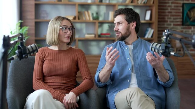 Webcam view. Man and woman talking using microphones while recording a podcast while sitting in a modern studio. Two smiling bloggers looking in camera, having a conversation, interview for video blog