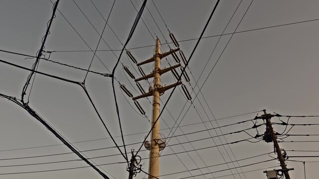 A tall steel utility pole with ceramic insulators rises through a dense tangle of overhead electric cables beneath a flat gray sky, capturing everyday urban infrastructure.