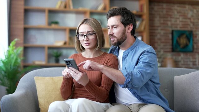 Married couple shopping choosing products in online store using smartphone sitting on sofa in living room at home. Man and woman buyers use online app store for discount, sale promotions on phone