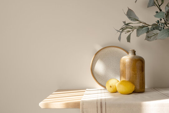Ceramic dishes, lemons on wooden table with beige linen runner, floral branch, empty warm beige wall background copy space, natural sunlight shadows. Cozy cottage Mediterranean kitchen interior