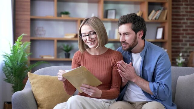 Happy married couple celebrating success after reading a letter with great news while sitting on sofa in living room at home. Excited smiling man and woman satisfied with pleasant good notification
