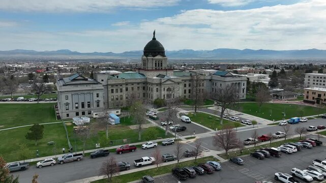 Montana State Capitol with copper dome rises above green lawns and full parking lots in Helena, with low-rise cityscape and Rocky Mountain foothills beyond. Montana, USA. Aerial drone approaching shot