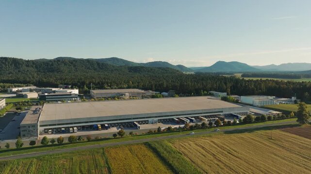 Large logistics center shows trucks loading and unloading cargo in the distribution area with mountains in the background and clear skies.