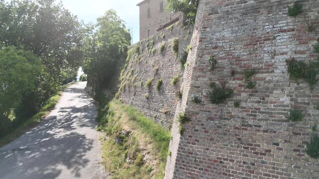 Strada stretta che costeggia un alto muro in mattoni con vegetazione spontanea, sormontato da edifici storici. Scena urbana antica immersa nel verde, luminosa e caratteristica