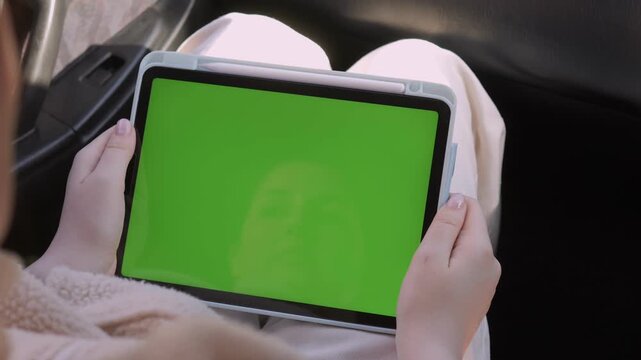 Person holds tablet with green screen while seated in car. Hands steady, focused on device. Ideal for digital media or advertising use. Suitable for travel, work, or leisure scenarios
