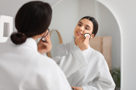 Young woman removing makeup with cotton pad near mirror in bathroom