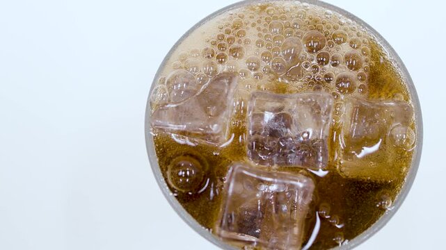 A top view close-up of cola with ice cubes and foam bubbling at the surface of a glass against a white background.