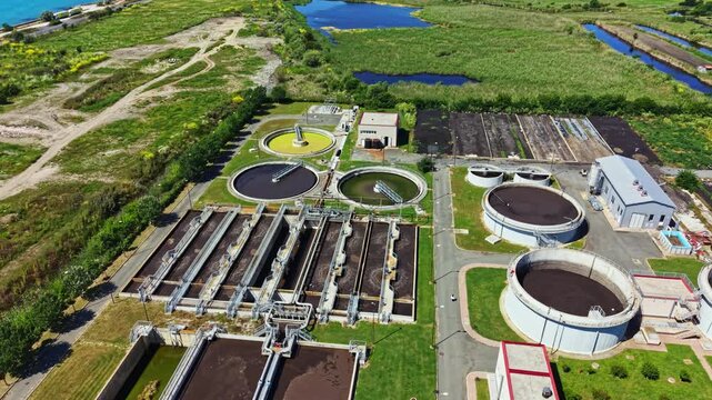 A water treatment facility with several large tanks and basins used for cleaning wastewater. The facility sits close to a river and green fields.