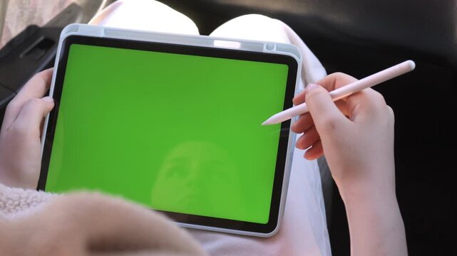 Person holds tablet with green screen. Hand uses stylus to draw or write. View shows focused creative work. Ideal for digital art or design projects. Suitable for advertising, tutorials