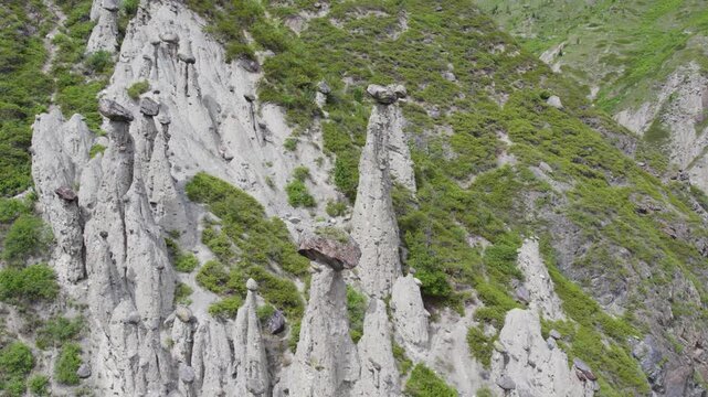 Aerial view of tall hoodoo rock pillars with balancing capstones on steep vegetated slope, showing weathered stone towers, layered cliff texture, remote mountain landscape, natural erosion forms and