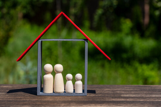 Minimalist wooden house model with red roof protecting small peg doll family on desk with blurred green nature background representing real estate and safety