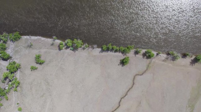 Aerial riverbank view showing reflective river surface beside dry pebble floodplain, shoreline structures, scattered vegetation, and winding drainage channel beneath bright daylight