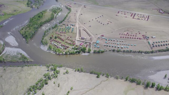 Oblique aerial view moving over riverside campsite with organized cabin and tent layout beside winding river and sandy floodplain under soft overcast light, with aerial, riverside, campsite