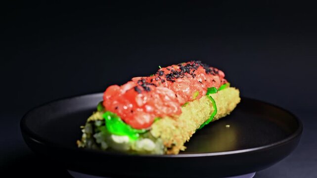Sushi roll is placed on a dark plate in a restaurant. The roll has layers of avocado and fresh fish with toppings. It is set in a simple environment, highlighting the food.