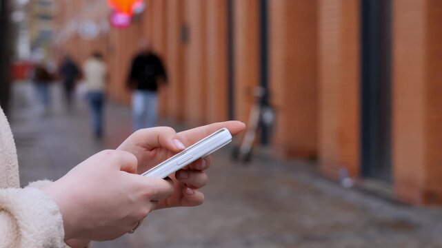 Person holds phone, focused on screen, walking down urban street. Background shows blurred pedestrians and brick walls. Suitable for themes of communication, daily commute, or mobile usage
