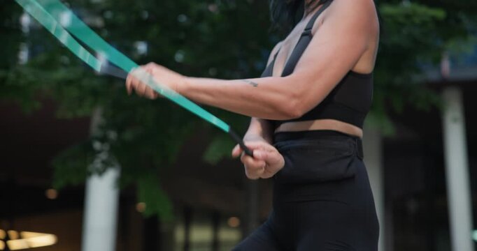 Young beautiful female athlete in sportswear training and jumping rope in an urban park among high-rise buildings and trees - hands close-up