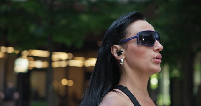 Young beautiful female athlete in sportswear trains in an urban park among high-rise buildings and trees and rests during a break between exercises