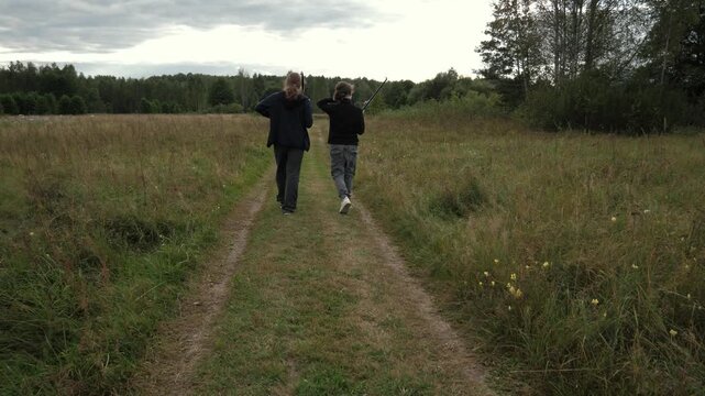 A rear view of two teenagers walking with fishing rods along a narrow dirt path through tall meadow grass toward mixed deciduous trees under an overcast sky. Soft, diffused daylight