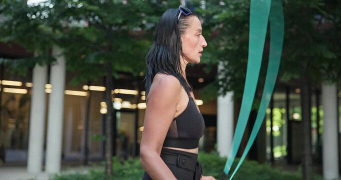 Young beautiful woman athlete in sportswear training and jumping rope in an urban park among high-rise buildings and trees