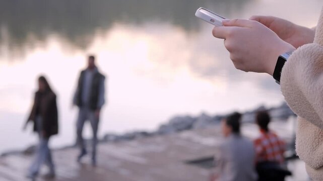 Close-up of hands holding a phone, capturing a blurred scene by water. Background shows people relaxing near a lake at sunset. Calm, peaceful mood with warm golden light. Ideal for social media