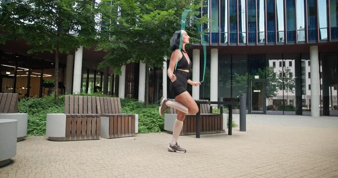 Young beautiful woman athlete in sportswear training and jumping rope in an urban park among high-rise buildings and trees