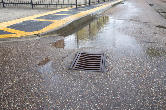 Puddle of water on wet asphalt pavement near a storm drain grate after rain. Urban street scene. Real Photo