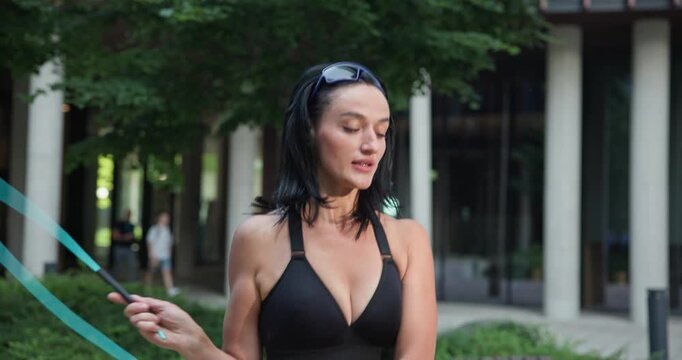 Young beautiful woman athlete in sportswear training and jumping rope in an urban park among high-rise buildings and trees