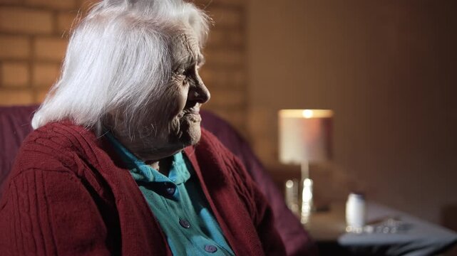 An elderly, gray-haired woman sits on sofa in a gloomy room, looking bewildered