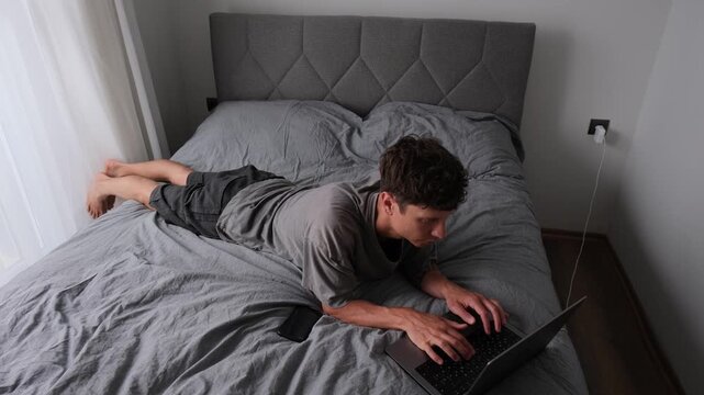 Young man adapting bedroom into workspace while working remotely on laptop, authentic freelance lifestyle in compact apartment with casual everyday atmosphere