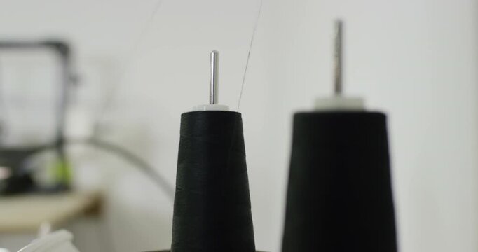 Close up of two black thread spools on holders in a sewing room with blurred equipment in background