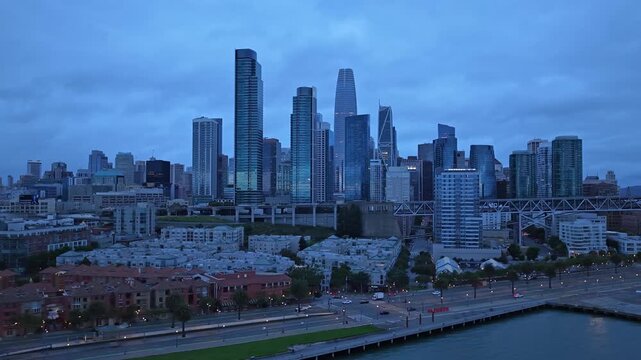 Panning drone view captures San Francisco&rsquo;s iconic skyline at dusk, featuring modern skyscrapers, moody blue hour lighting, waterfront, and dramatic cityscape composition.