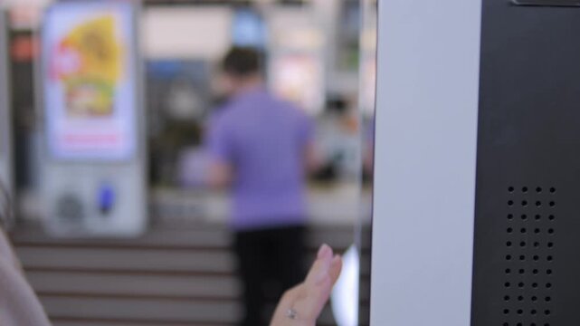 Person's hand gently touches a modern kiosk screen. The background shows a blurred indoor setting with another person moving. The focus stays on the interaction, highlighting touch technology use