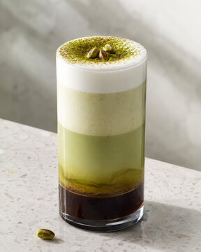 Layered Pistachio Latte in Clear Glass with Vibrant Green Pistachio Powder and Whole Pistachios on White Foam, Terrazzo Surface with Dappled Shadow, Specialty Coffee Art