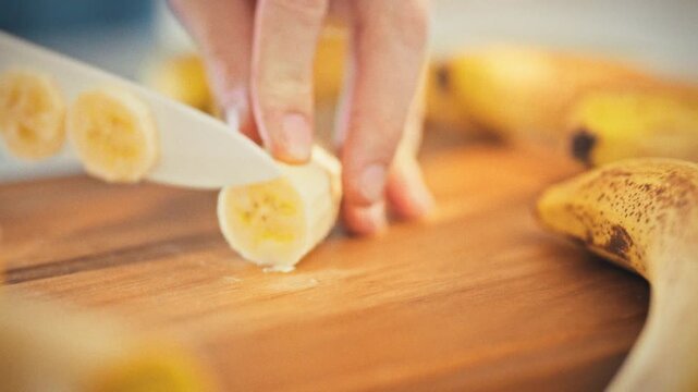 In a warm home kitchen bathed in sunny light, a man's hands carefully slice a fresh banana with a ceramic knife on a wooden cutting board, embodying the cozy essence of cooking and living at home
