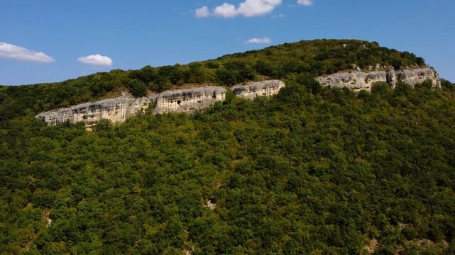 Aerial slow motion pan of forested hill revealing exposed limestone cliff on sunny summer day