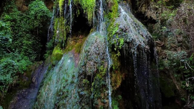 Panning shot of waterfall cascading over moss covered rocks with lush foliage in shaded forest gorge during daytime natural scene