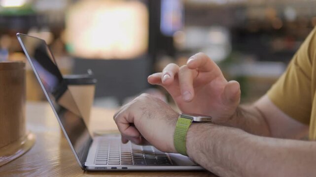 Close-up of hands typing on laptop in cozy cafe. Person wears green watch, focused on screen. Background shows warm lighting and blurred interior. Ideal for remote work, online meetings
