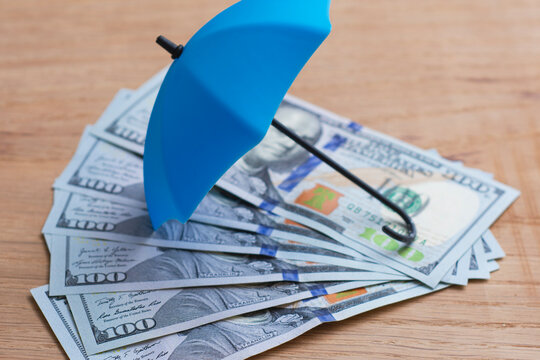 US dollar banknotes protected by a blue umbrella on a wooden surface. Financial security, money protection, insurance coverage, wealth preservation, investment safety and risk management concept.