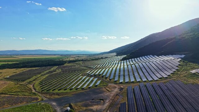 Solar panels are arranged in rows across a field at the foot of a mountain. The sun shines down on the scene as clouds drift by. The area is dedicated to producing renewable energy.
