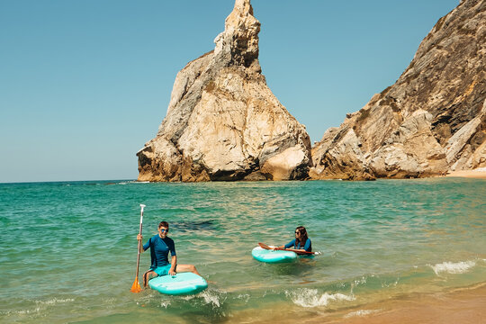 beach Ursa, Portugal. Two individuals are paddleboarding on a beach with clear blue water. They are close to rocky cliffs under a bright sky. Other people swim in the background.