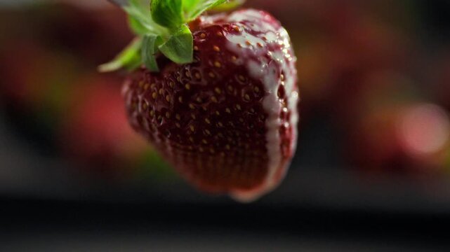A close-up of white whipped cream slowly dripping down a ripe red strawberry. The dark, blurred background of berries creates a rich depth in the frame. Concept for a fresh summer dessert