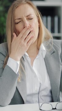 Woman in a gray suit looks sleepy while working in an office. She yawns and shows fatigue.