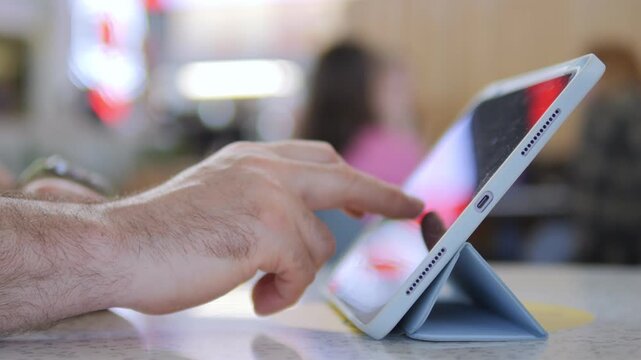 Hand taps tablet screen in cafe setting. Fingers interact with digital display. Background shows blurred people. Suitable for tech, communication, or work-from-anywhere themes