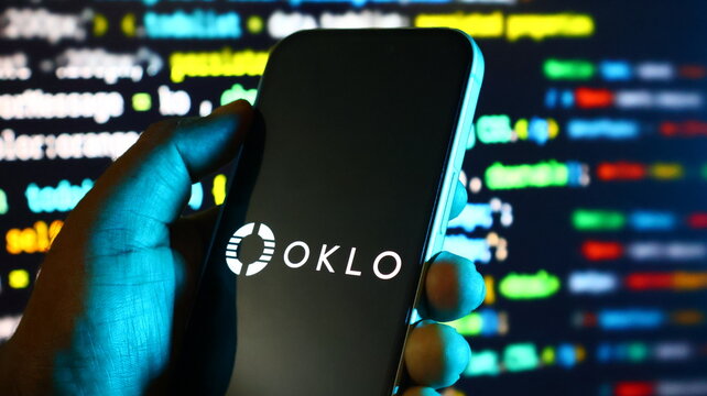 Smartphone displaying oklo logo held in hand with computer code background in night light.