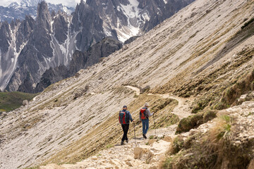 Fototapeta premium Two hikers with backpacks and trekking poles walking on a rocky mountain trail beneath jagged peaks, alpine hiking adventure concept 