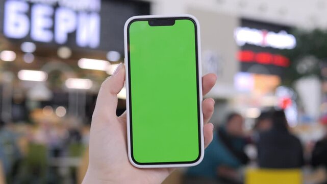 Hand holds phone with green screen. Background shows busy indoor market. Blurry people move around. Ideal for app mockups or advertising. Suitable for retail, tech, or digital marketing use