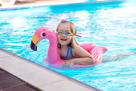 Happy little girl wearing swimming goggles floating on a pink flamingo inflatable in a blue pool. Fun summer vacation, water safety, childhood joy, and family leisure concept