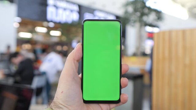 Hand holds smartphone with green screen. Background shows blurred indoor public space. People move behind phone, unaware of camera. Ideal for app mockups, digital marketing, or UI design projects