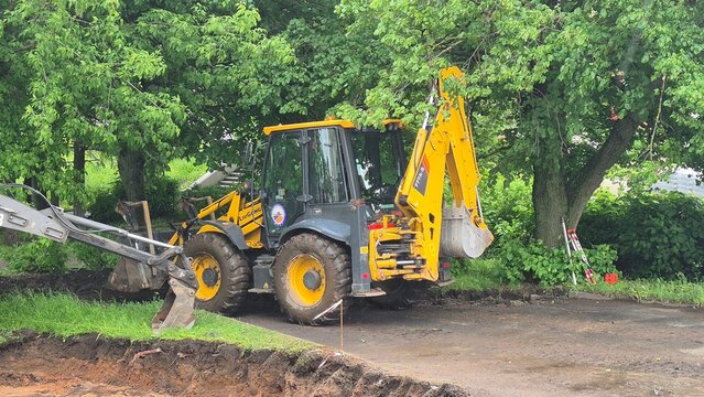 Backhoe loader performing road maintenance work beside a park in an urban area. Municipal infrastructure maintenance and public utility operations. Rybinsk, Russia, May 27, 2026.